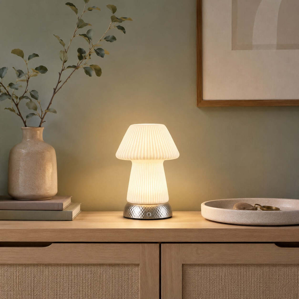 A glowing Clérachic lamp on a rattan-fronted oak cabinet, with a stone vase of olive stems, a framed print and a shallow dish nearby.