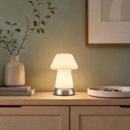 A glowing Clérachic lamp on a rattan-fronted oak cabinet, with a stone vase of olive stems, a framed print and a shallow dish nearby.
