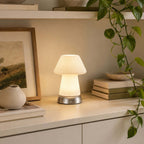 Close beside a framed landscape and a trailing green plant, this Clérachic fluted lamp lights a pale shelf with stacked books and a vase.