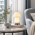 Softly lit on a marble side table by a bright window, this Clérachic fluted lamp stands near a bowl, books and a blue armchair.