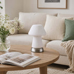 Unlit, this white fluted Clérachic lamp stands on a wooden coffee table before a cream sofa, with a magazine and a jar of white flowers.