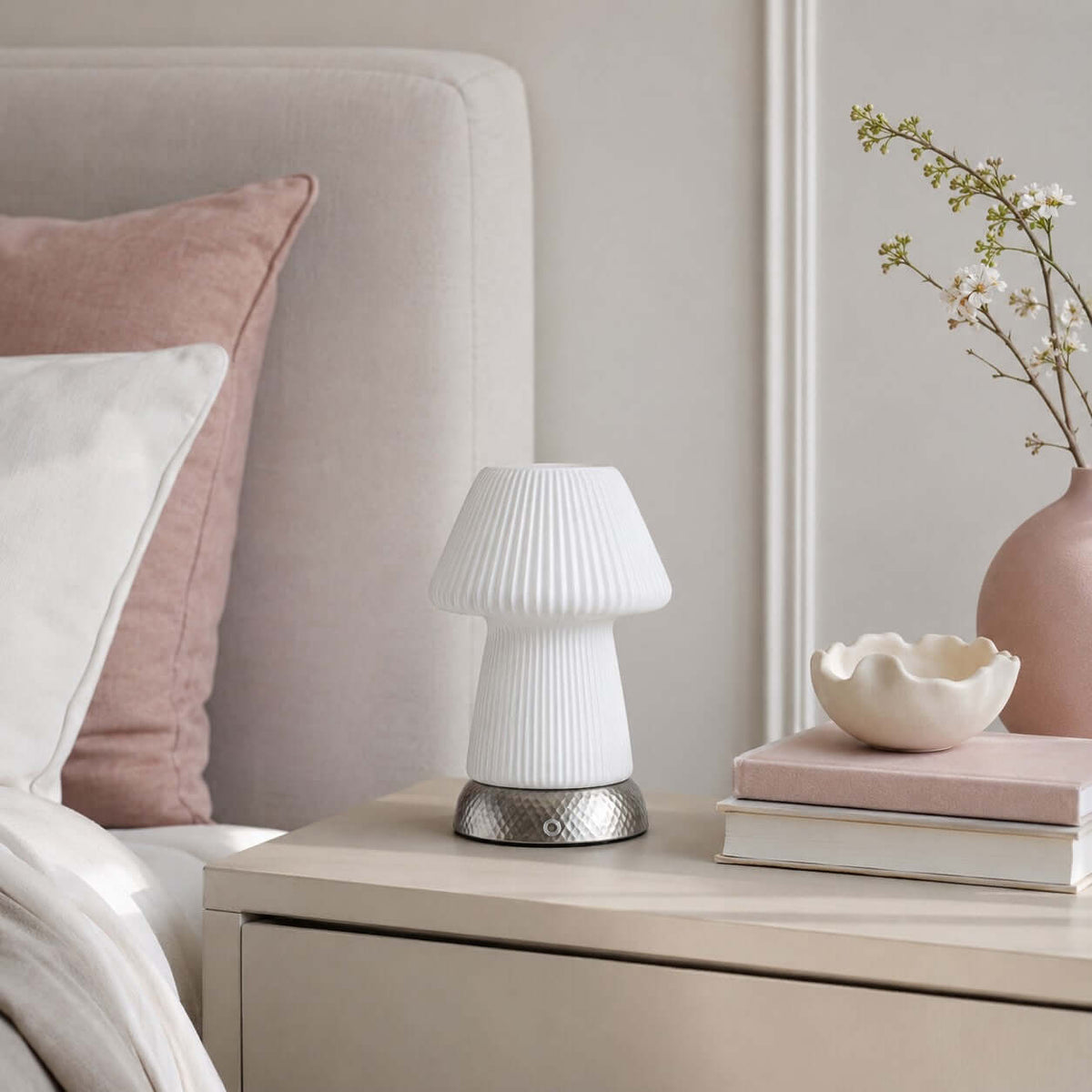 On a pale bedside chest, this unlit white Clérachic lamp stands near pink cushions, a shell-shaped dish, a stack of books and flowering stems.