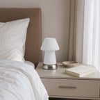 Beside a rumpled white bed and a taupe headboard, this Clérachic fluted lamp sits unlit on a bedside chest with a book and a small dish.