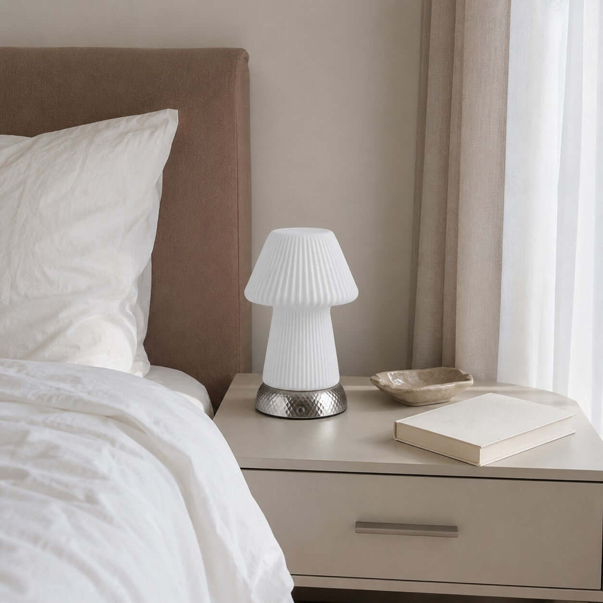 Beside a rumpled white bed and a taupe headboard, this Clérachic fluted lamp sits unlit on a bedside chest with a book and a small dish.
