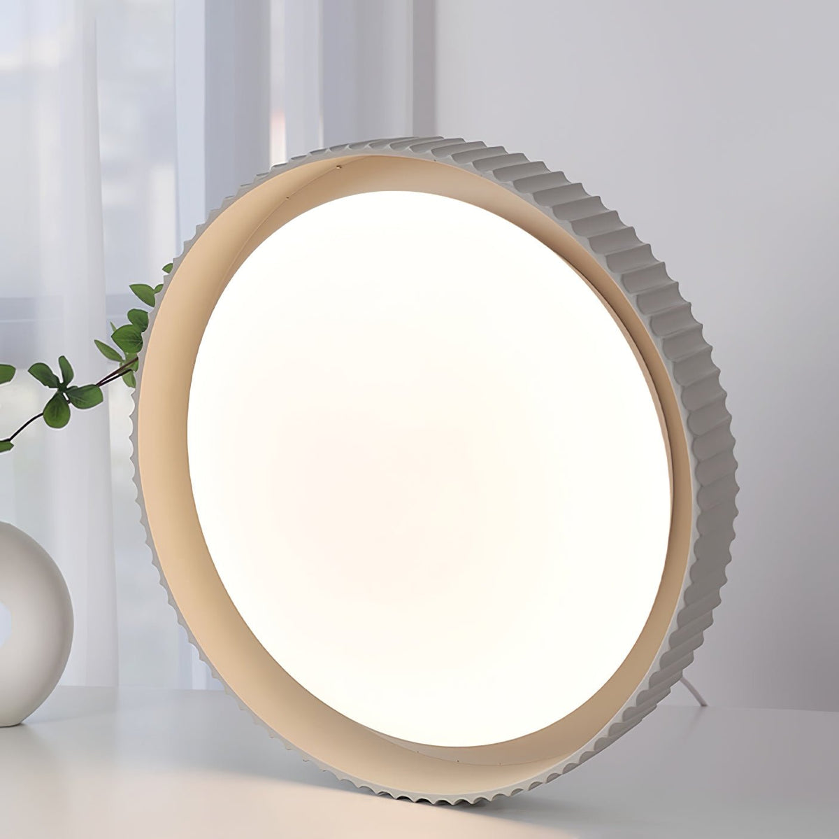 A Clérachic ribbed flush light stood on its edge on a white surface beside a plant, its large white diffuser facing the viewer.