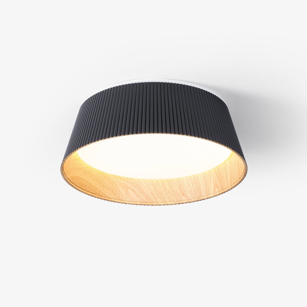 A black fluted Clérachic ceiling light with a wood-grain inner rim and a glowing diffuser, seen from below on a white backdrop.