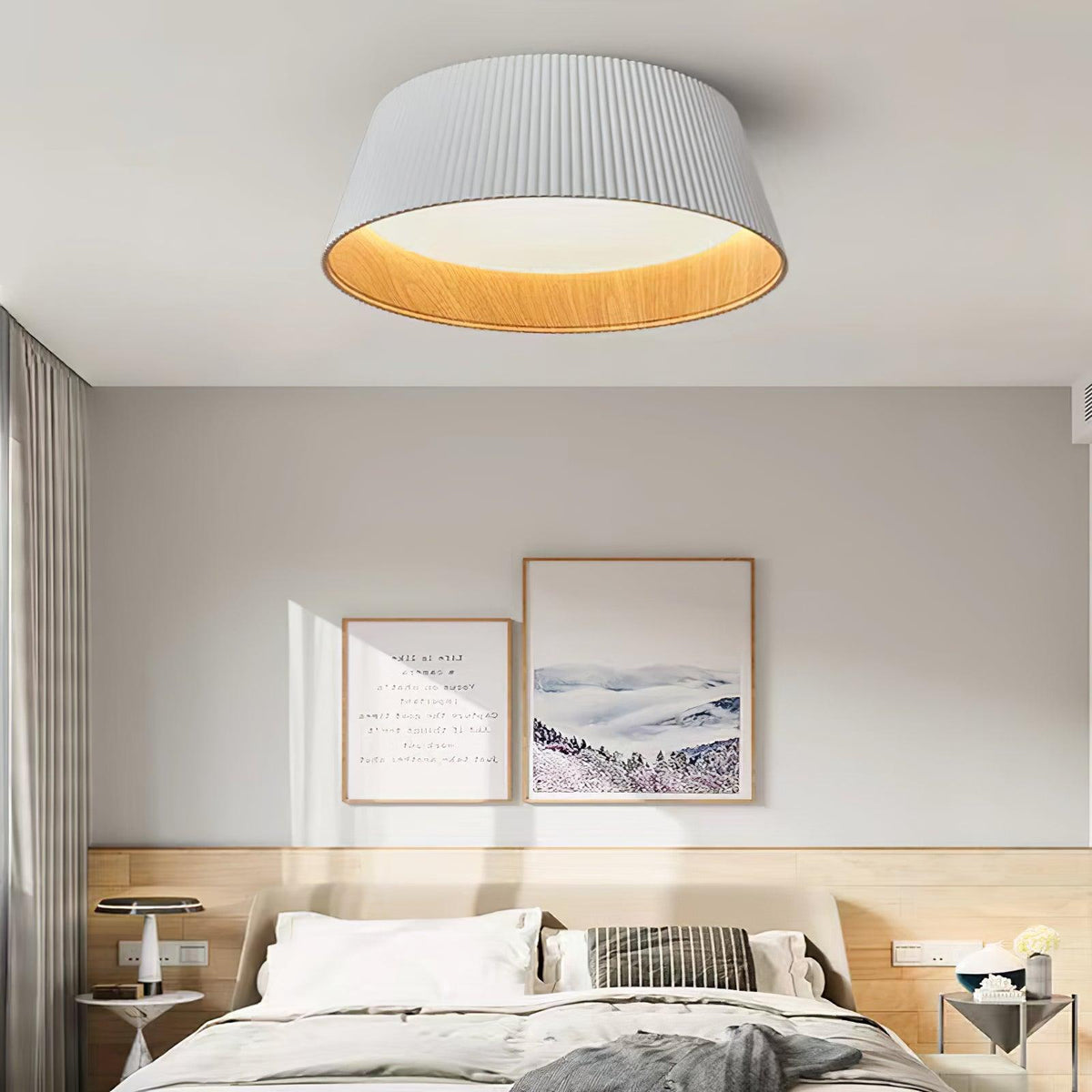 A white fluted Clérachic ceiling light above a bed with a pale timber headboard, with two framed prints on a grey wall.