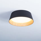 A lit black Clérachic ceiling light with a warm glowing diffuser and a wood-grain inner band, on a pale blue-grey ceiling.