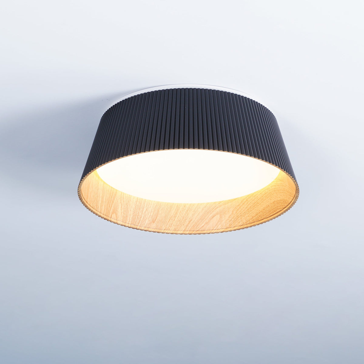 A lit black Clérachic ceiling light with a warm glowing diffuser and a wood-grain inner band, on a pale blue-grey ceiling.