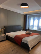 Black Clérachic ceiling light at the top of a bedroom with a dark leather bed, a red throw and a window with sheer curtains.