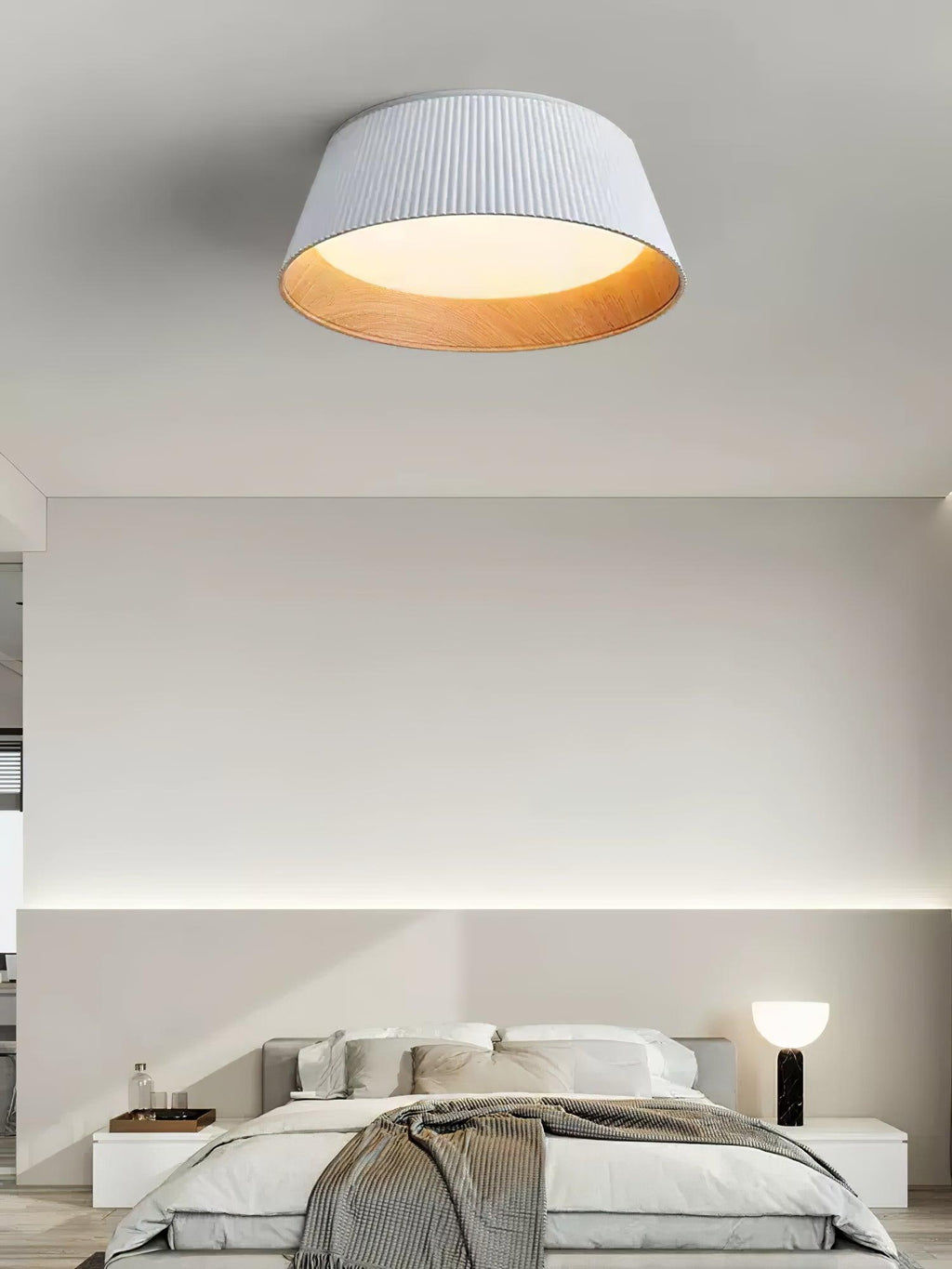 A white Clérachic ceiling light in a minimal bedroom, with a black table lamp on the bedside and a cove light behind.