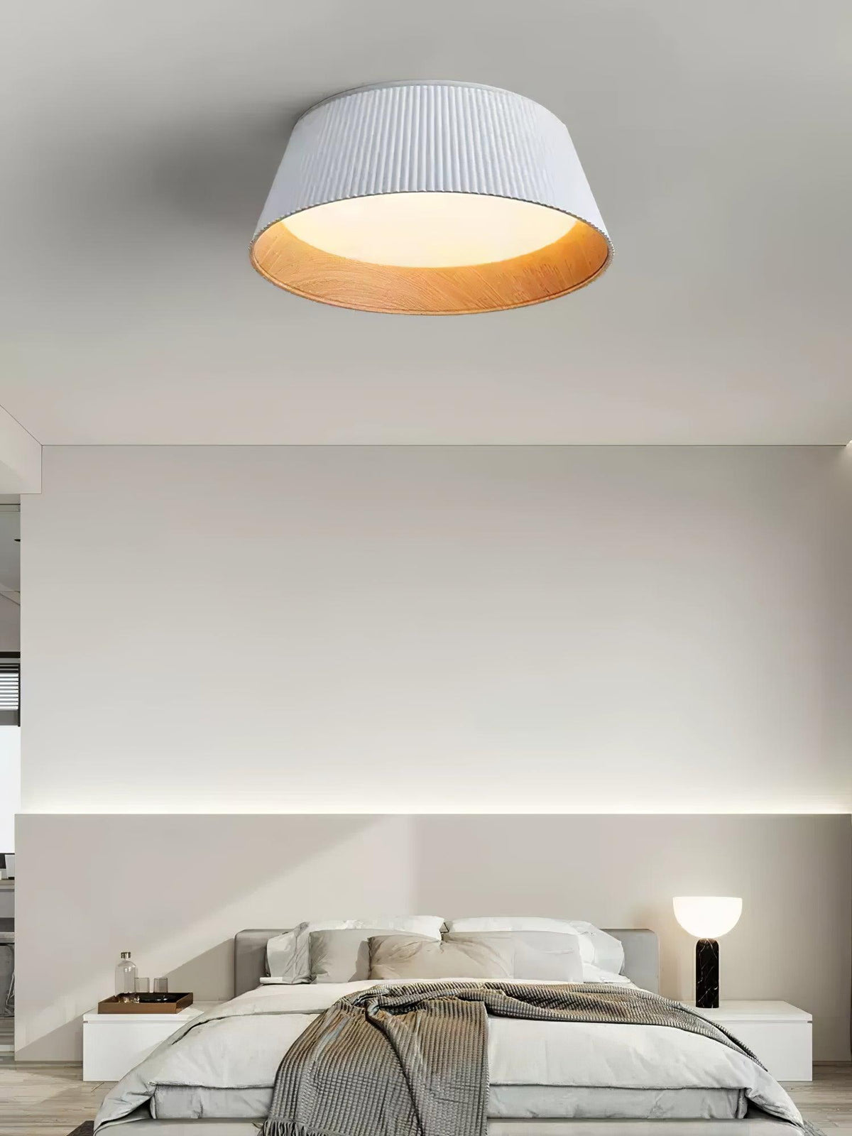 A white Clérachic ceiling light in a minimal bedroom, with a black table lamp on the bedside and a cove light behind.