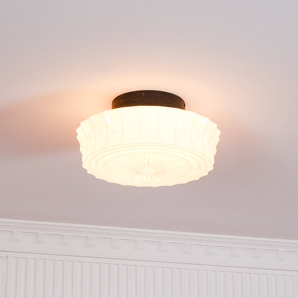 Close view beneath ornate crown molding of the white Clérachic fluted opal glass flush ceiling light, lit.
