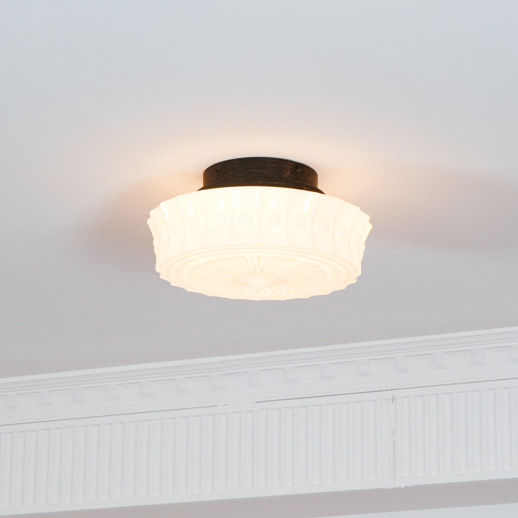 The white Clérachic fluted opal glass flush ceiling light lit in a room with plain walls and crown molding.