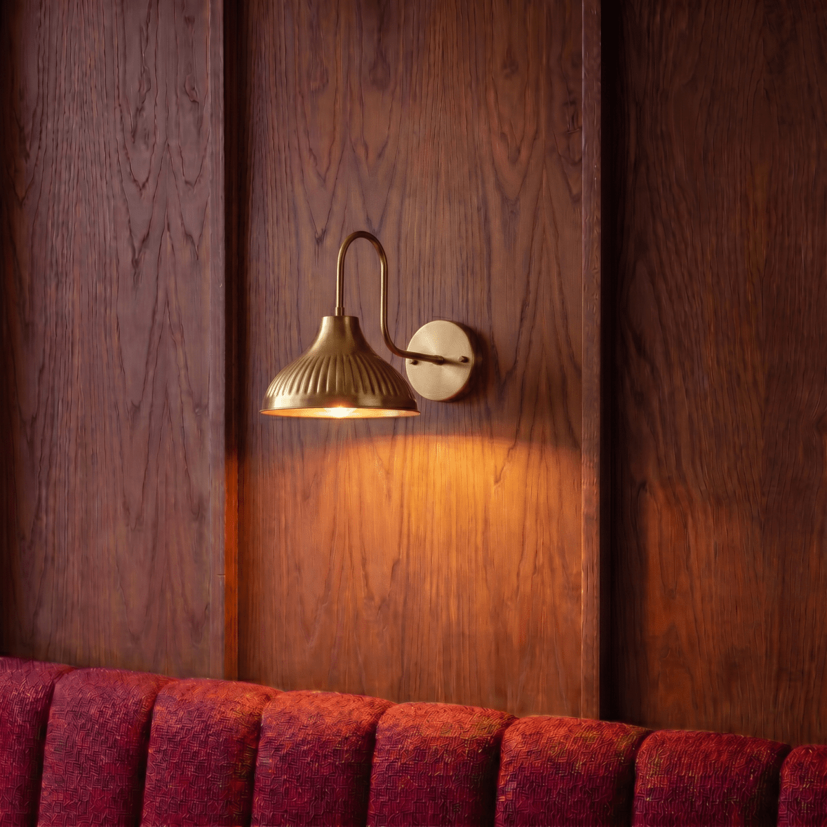 This brass Clérachic wall lamp lights a wood-paneled wall above a burgundy velvet banquette.