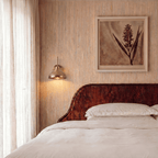 A vintage brass Clérachic wall lamp lights a bedroom headboard beneath a framed botanical print.