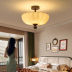 Lit scalloped cream shade of this Clérachic ceiling light above a bed, with green curtains and a woman at the window.
