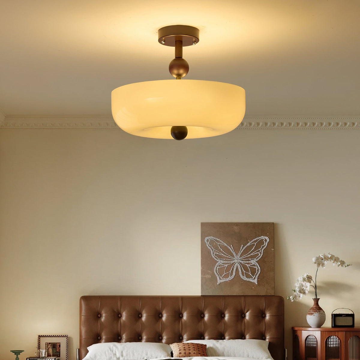 Glowing round cream drum of this Clérachic ceiling light over a tufted brown leather bed, with a butterfly artwork behind.