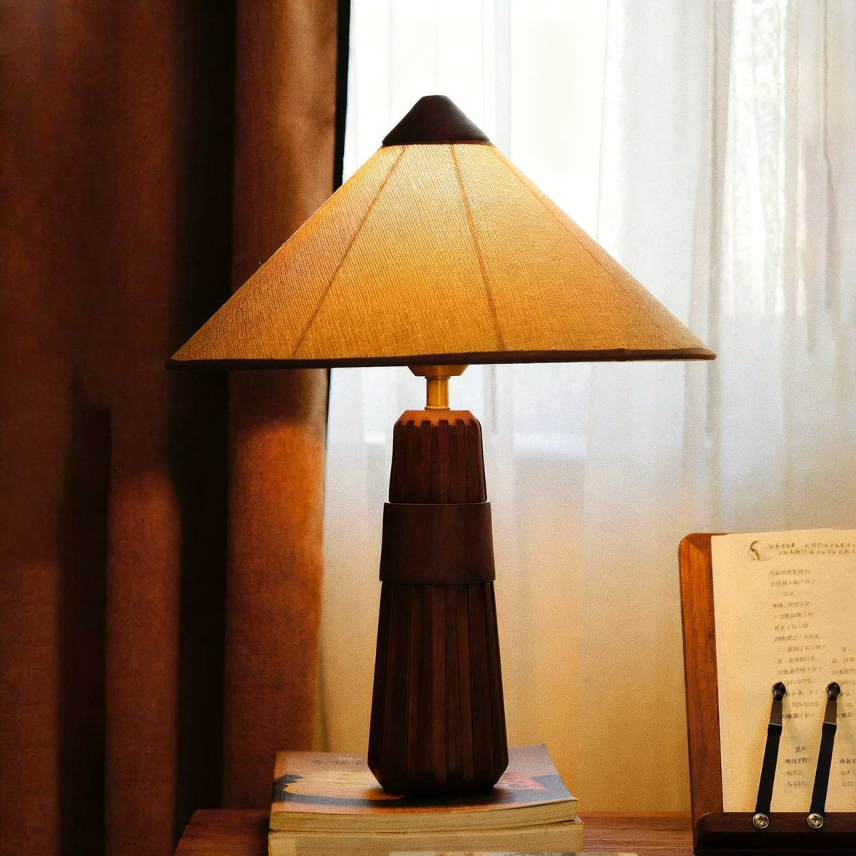Walnut-finish Clérachic table lamp with a warm-lit cone shade, on a pile of books by a rust curtain and an open book stand.