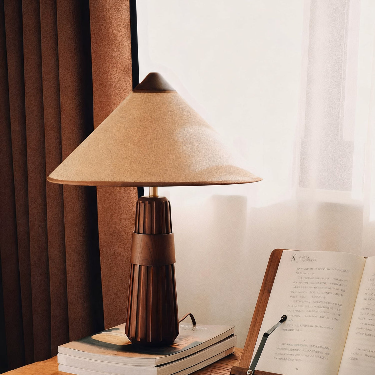 Beige-shaded Clérachic table lamp with a walnut base on stacked books, beside an open book and a sheer curtain.