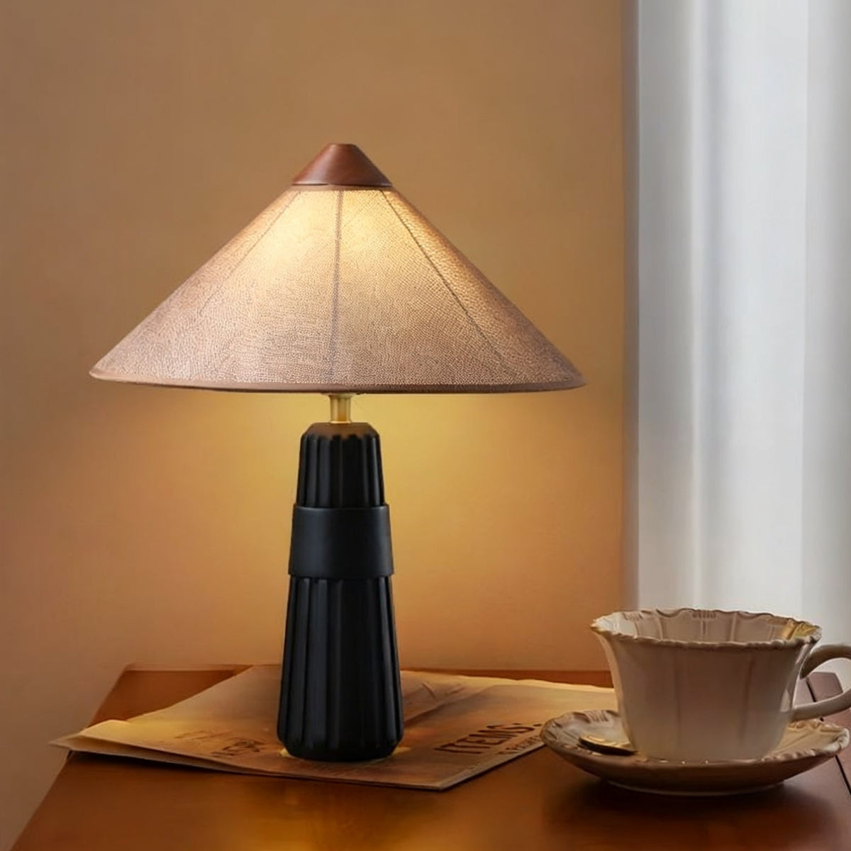 Black-base Clérachic table lamp with a soft pink-beige cone shade, beside a cup and saucer on a wooden bedside table.