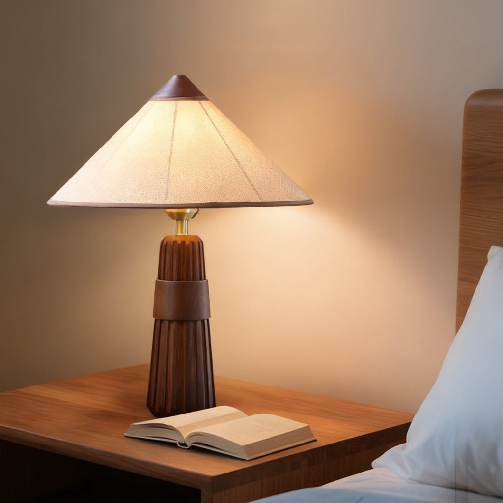 Walnut Clérachic table lamp lit on a wooden nightstand beside an open book, with a bed and white pillow at the edge.