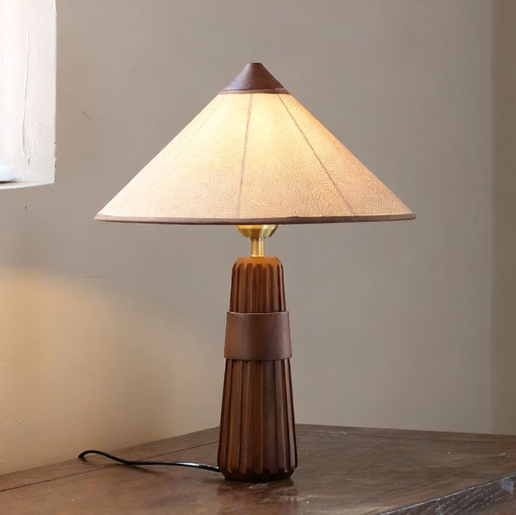 Walnut-base Clérachic table lamp with its cone shade glowing, on a dark wood surface with the cord trailing to the left.