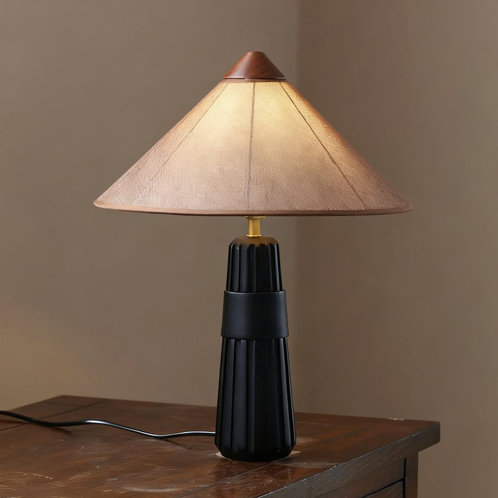Black fluted-base Clérachic table lamp with a muted pink-beige cone shade, on a dark wooden surface beside a window.