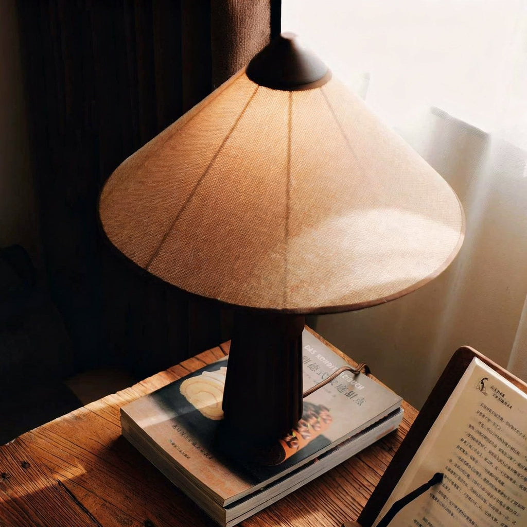Angled view down onto the linen-look cone shade of this Clérachic table lamp, on a stack of magazines by a curtain.
