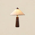 Walnut-base Clérachic table lamp with a cone shade, lit and shown against a plain warm cream backdrop.