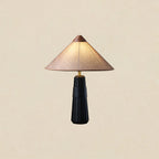 Black-base Clérachic table lamp with a pink-beige cone shade and wooden cap, lit against a plain warm cream backdrop.