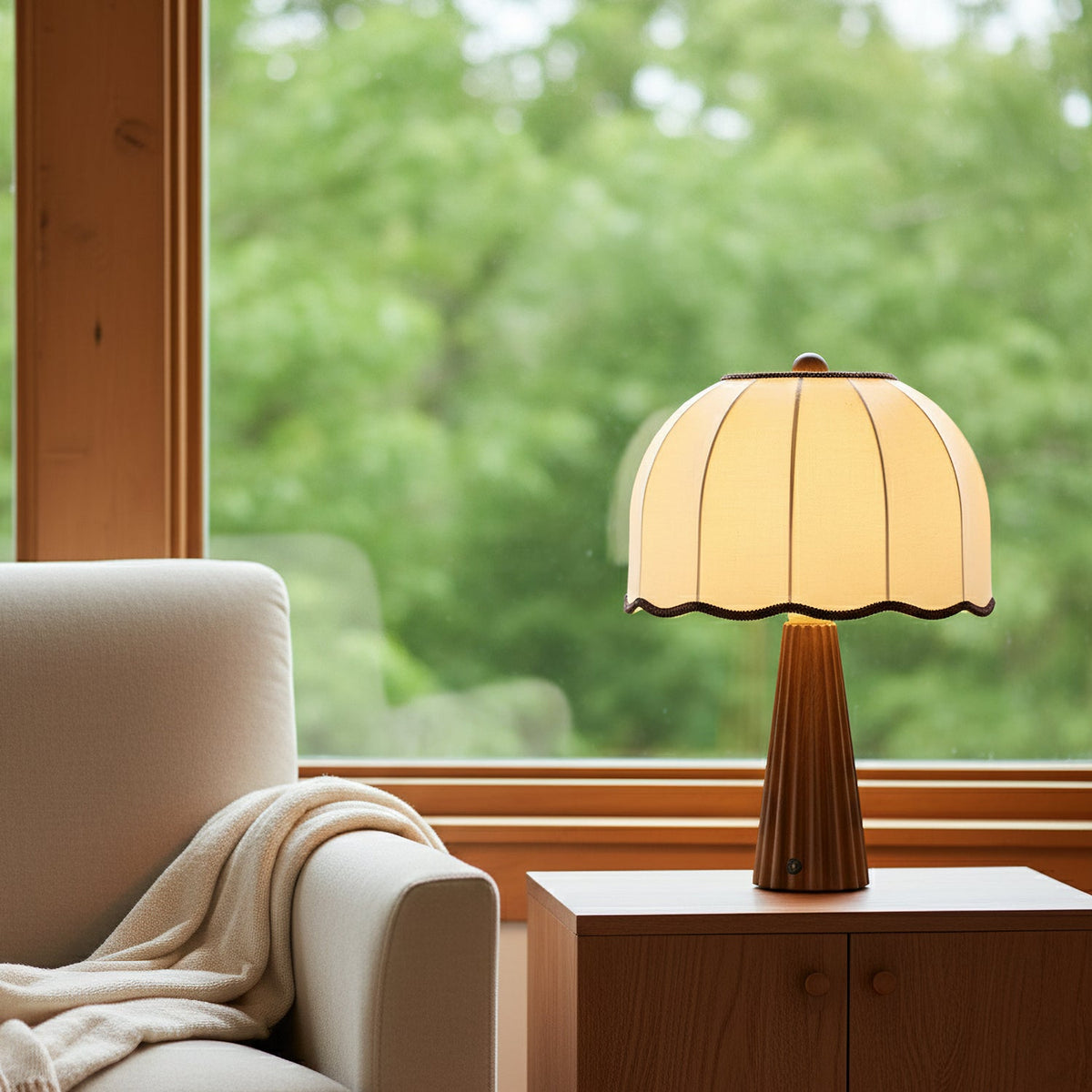 Clérachic dome-shaded table lamp on a wooden cabinet beside a window overlooking green trees, with a cream throw over a chair.