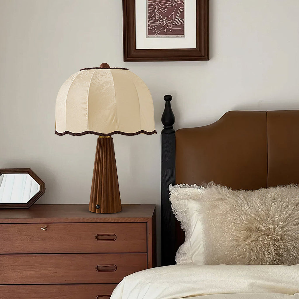 A Clérachic table lamp with a cream scalloped shade on a chest of drawers beside a bed with a tan leather headboard.