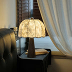 Lit Clérachic table lamp with a bamboo-print shade on a black bedside cabinet, with a sheer curtain and a dark leather headboard.
