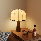 Lit Clérachic table lamp with a cream dome shade on a wooden side table with a bowl of nuts and a glass of whisky beside it.