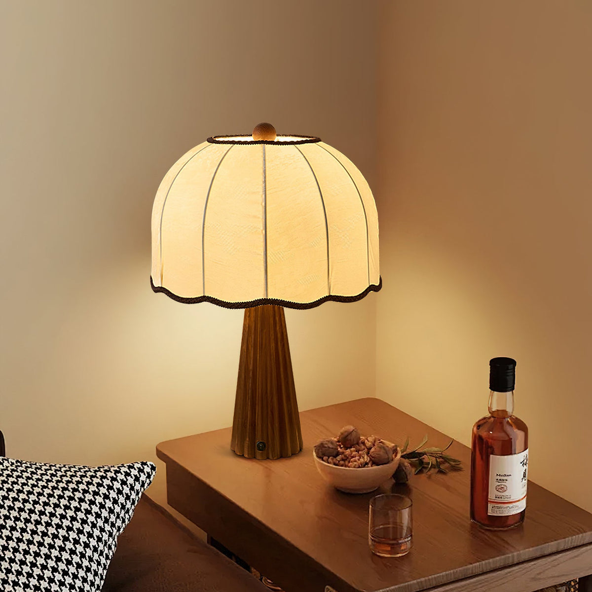 Lit Clérachic table lamp with a cream dome shade on a wooden side table with a bowl of nuts and a glass of whisky beside it.
