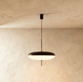 Clérachic flying-saucer pendant with a matt black top and glowing white acrylic underside on a thin black rod, against limewash.