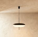 Clérachic flying-saucer pendant with a matt black top and glowing white acrylic underside on a thin black rod, against limewash.
