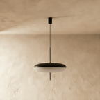 Clérachic saucer pendant in black and white, hung on a slim black stem from a round black canopy against a pale plaster wall.