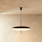 Clérachic wide black and white saucer pendant with a glowing white underside, on a thin black stem against a limewash wall.