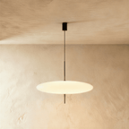 Clérachic wide all-white saucer pendant with a smooth opal diffuser on a slim black rod, lit against a textured plaster wall.