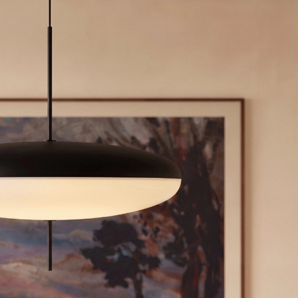 Clérachic close view of a black-topped saucer pendant with a warm white diffuser and a slim black rod in front of a framed painting.