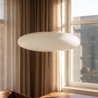 Clérachic close view of an all-white glass-like saucer pendant on a black rod, beside linen curtains and a bright window.