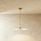 Clérachic frosted disc pendant with gold cone cap, gold centre panel and gold cord, against warm limewash plaster.