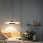 Clérachic black-centred disc pendant glowing above a bedside shelf with a plant, pillows and books in a grey bedroom.