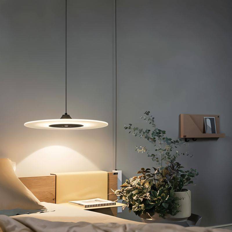 Clérachic black-centred disc pendant glowing above a bedside shelf with a plant, pillows and books in a grey bedroom.