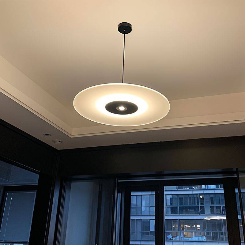 Clérachic black-centred disc pendant lit warm below a recessed ceiling, seen from beneath in a room with large windows.