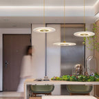 Clérachic three gold-centred disc pendants at different heights above a pale wooden table with green stools.
