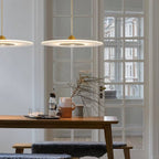 Clérachic gold-capped disc pendants glowing over a wooden dining table and bench beside tall white-framed glazed doors.