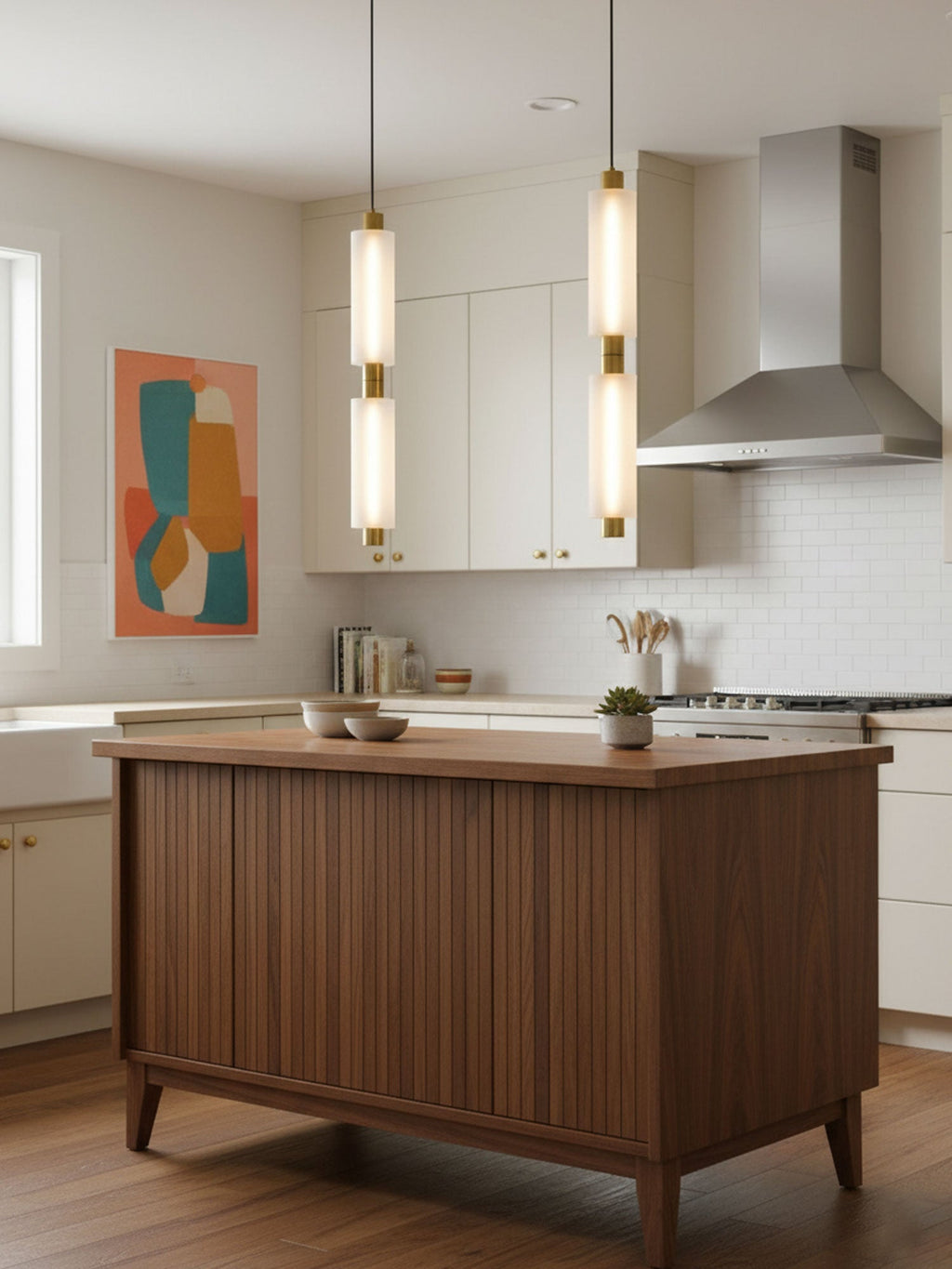 Two tall Clérachic pendants with gold joints hang in a white kitchen, beside a walnut ribbed island and a colourful print.